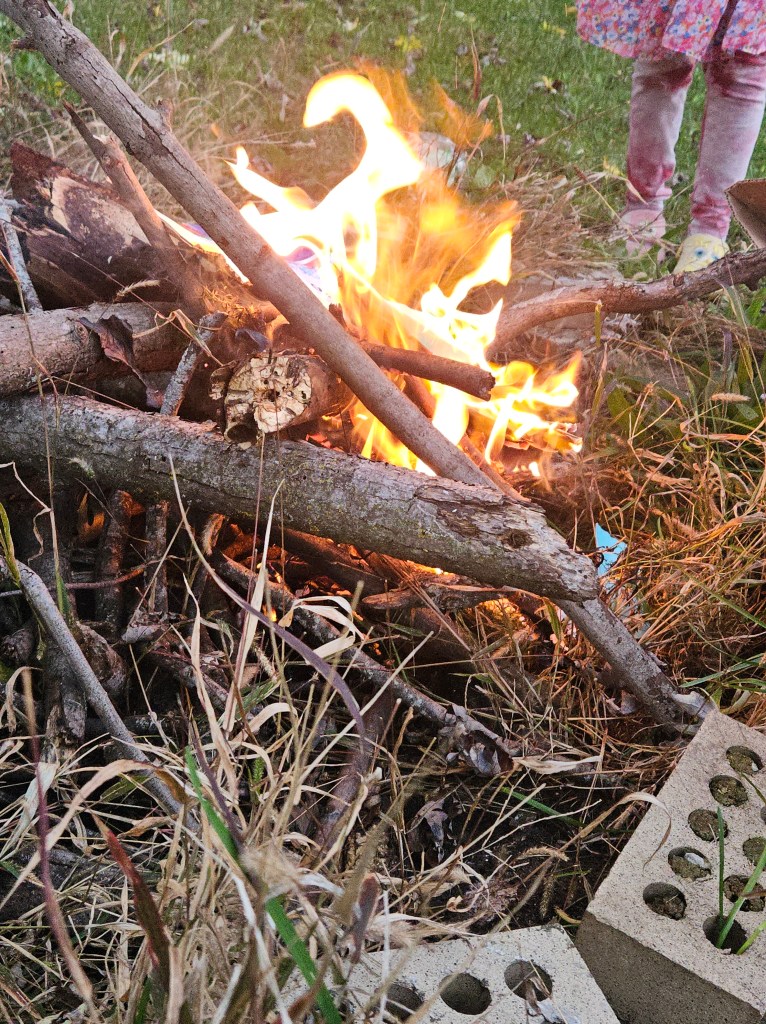 Photograph of a bonfire surrounded by cinder blocks and a small child's legs standing in the background. 