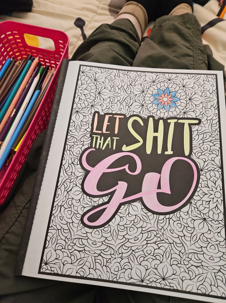 An opened coloring book page that says "Let that shit go". Partially colored with a basket of colored pencils to the left. The book rests on a lap. 