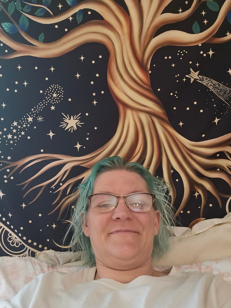 A woman wearing a white shirt, glasses, and teal hair smiles at the camera. A large brown tree tapestry is in the background with shooting stars near the tree. 